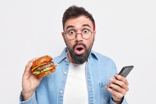 Foto gratuita hombre adulto barbudo hambriento come deliciosa hamburguesa sostiene teléfono móvil descubre noticias impactantes lleva camisa de mezclilla de gafas redondas