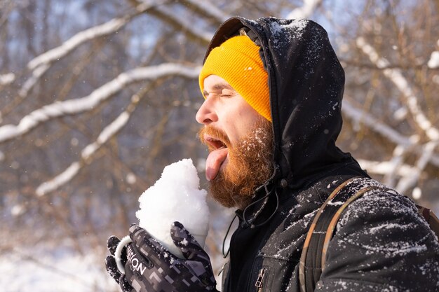 Hombre adulto brutal en un sombrero naranja brillante en un bosque nevado en un día soleado con una taza llena de nieve divirtiéndose