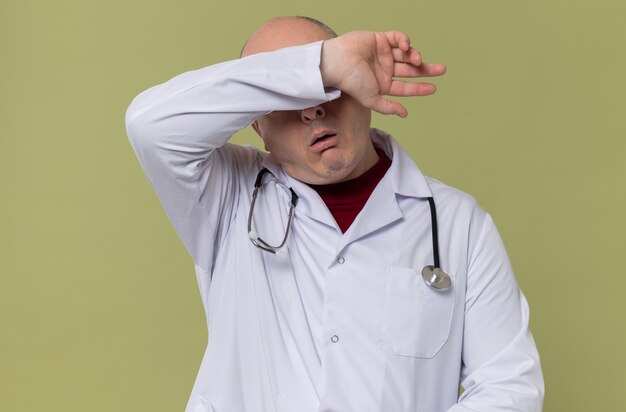 Hombre adulto cansado en uniforme médico con estetoscopio poniendo el brazo en la frente