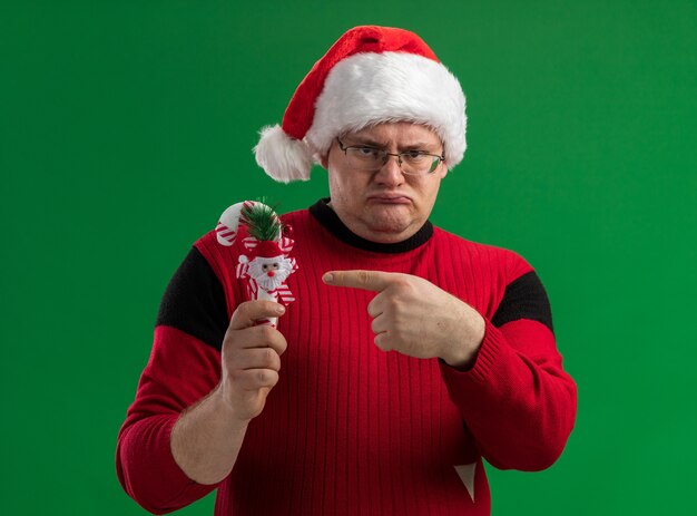 Hombre adulto con el ceño fruncido con gafas y gorro de Papá Noel sosteniendo apuntando al adorno de bastón de caramelo mirando a cámara aislada sobre fondo verde