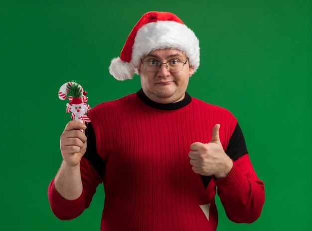 Hombre adulto complacido con gafas y gorro de Papá Noel con adorno de bastón de caramelo mostrando el pulgar hacia arriba mirando a la cámara aislada sobre fondo verde