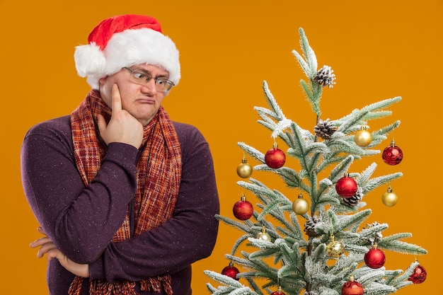 Hombre adulto confundido con gafas y gorro de Papá Noel con bufanda alrededor del cuello de pie cerca del árbol de Navidad decorado mirando hacia abajo manteniendo la mano en la barbilla aislada en la pared naranja