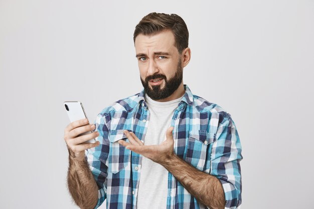 Hombre adulto decepcionado confundido reaccionando a una extraña aplicación en el teléfono inteligente