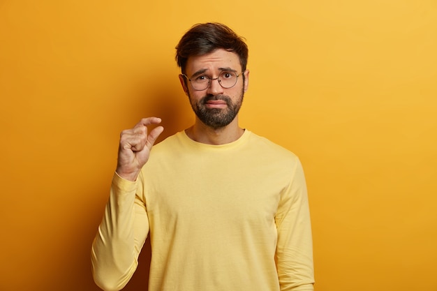 Un hombre adulto decepcionado muestra un tamaño pequeño, mide algo muy pequeño con los dedos, muestra una longitud o grosor insuficientes, discute precios reducidos, se viste con ropa informal, posa en interiores