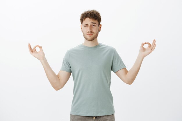 Hombre adulto divertido desenfocado con cerdas en aretes, mirando con un ojo mientras está de pie con las manos extendidas en gesto zen y practicando yoga o meditación