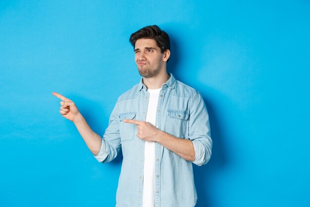 Hombre adulto dudoso señalando con el dedo a la izquierda en la promoción y mirando inseguro, haciendo muecas decepcionado, de pie contra el fondo azul