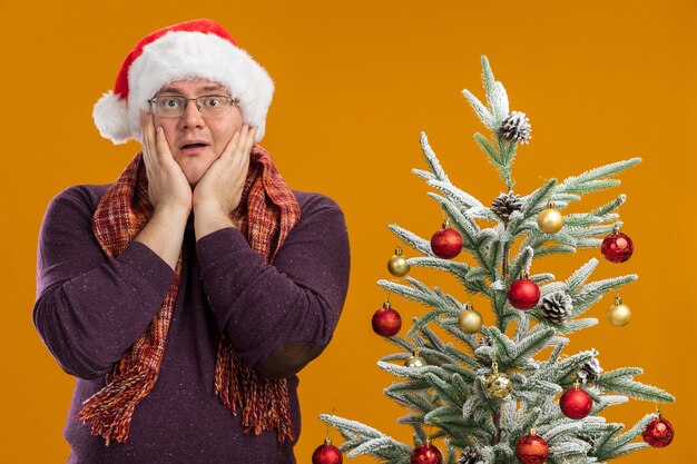 Hombre adulto emocionado con gafas y gorro de Papá Noel con bufanda alrededor del cuello de pie cerca del árbol de Navidad decorado manteniendo las manos en la cara mirando a la cámara aislada sobre fondo naranja