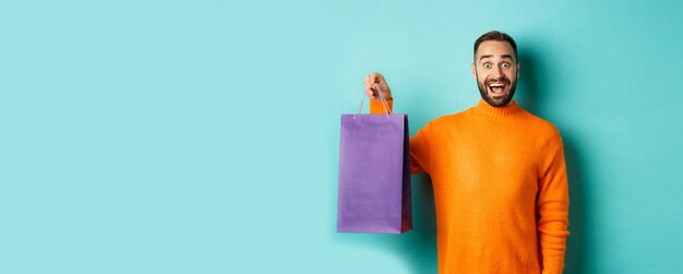 Foto gratuita hombre adulto emocionado sosteniendo una bolsa de compras morada y sonriendo yendo al centro comercial de pie sobre la espalda turquesa