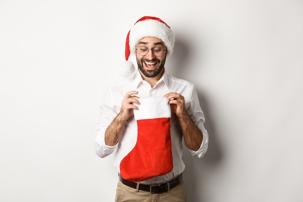 Hombre adulto feliz abra el calcetín de Navidad y mirando hacia adentro, recibiendo regalos de Navidad para las vacaciones de invierno, de pie con gorro de Papá Noel