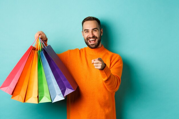 Hombre adulto feliz apuntando con el dedo a la cámara, sosteniendo bolsas de la compra y sonriendo