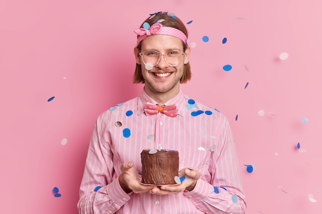 El hombre adulto feliz celebra un año de trabajo en compañía sostiene un pequeño pastel que recibe la felicitación de sus colegas sonríe con alegría viste una camisa a rayas con diadema y una pajarita cofetti cayendo sobre él