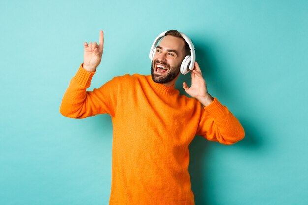 Hombre adulto feliz en suéter naranja, mirando hacia arriba y escuchando música en auriculares de pie contra la pared turquesa