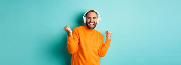 Hombre adulto feliz en suéter naranja mirando hacia arriba y escuchando música en auriculares de pie sobre ba azul