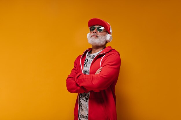 Hombre adulto con gorra roja y capucha escuchando música en auriculares sobre fondo naranja Chico elegante con barba blanca en gafas de sol azules posando