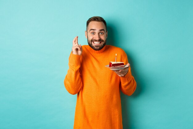 Hombre adulto guapo celebrando un cumpleaños, soplando velas en el pastel y pidiendo deseos, de pie contra la pared turquesa