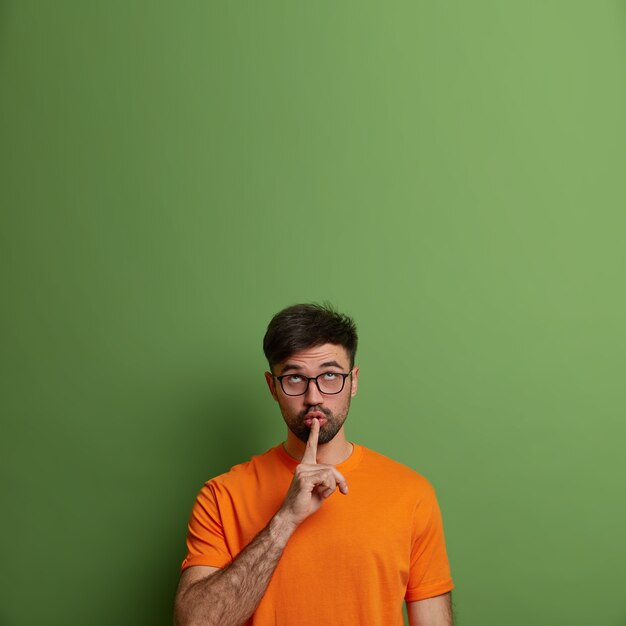 Hombre adulto guapo pide silencio, pone el dedo en la boca, se concentra arriba, dice información secreta, hace un gesto de silencio, vestido con una camiseta naranja informal, aislado en la pared verde