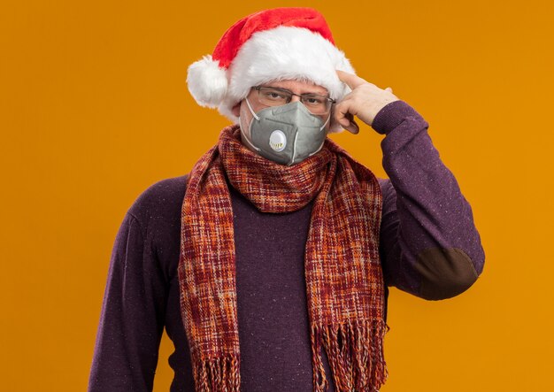Hombre adulto harto con gafas de máscara protectora y gorro de Papá Noel con bufanda alrededor del cuello mirando a la cámara haciendo gesto de suicidio aislado sobre fondo naranja