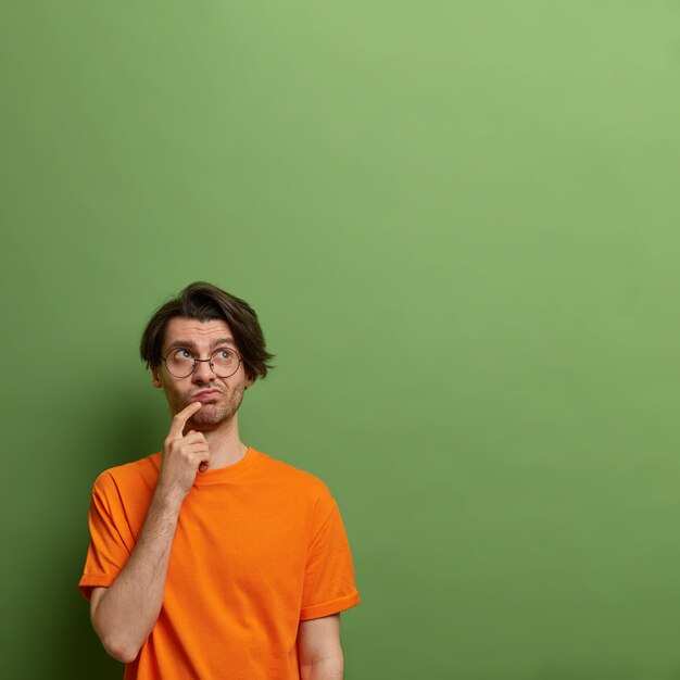 Hombre adulto indeciso pensativo mira hacia arriba y mantiene el dedo cerca de la boca, vestido con una camiseta naranja informal, posa contra la pared verde con espacio de copia para su promoción