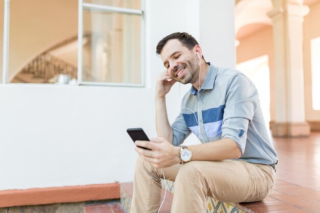 Hombre adulto medio feliz que usa la aplicación para escuchar música en el teléfono inteligente mientras está sentado en la entrada