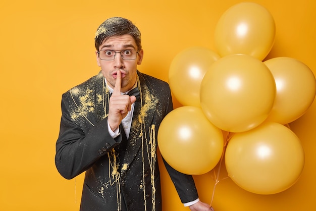 Hombre adulto misterioso sorprendido hace gesto de silencio usa anteojos traje formal negro manchado con spray serpentino sostiene un montón de globos inflados disfruta de la celebración de la fiesta Silencio, silencio