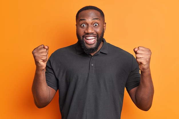 El hombre adulto negro positivo hace que el gesto de sí apriete los puños se siente como el campeón o el ganador viste una camiseta negra casual aislada sobre una pared naranja vívida