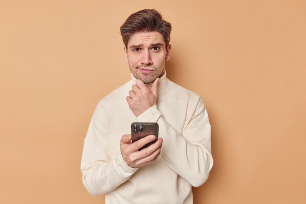 Hombre adulto pensativo desconcertado sostiene la barbilla y reflexiona sobre algo que sostiene el teléfono móvil considera la notificación recibida piensa qué respuesta dar usa cuello alto aislado en la pared beige del estudio