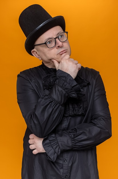 Hombre adulto pensativo con sombrero de copa y gafas en camisa gótica negra sosteniendo su barbilla y mirando a un lado