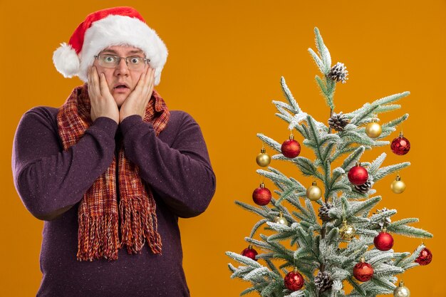 Hombre adulto preocupado con gafas y gorro de Papá Noel con bufanda alrededor del cuello de pie cerca del árbol de Navidad decorado manteniendo las manos en la cara aislada en la pared naranja