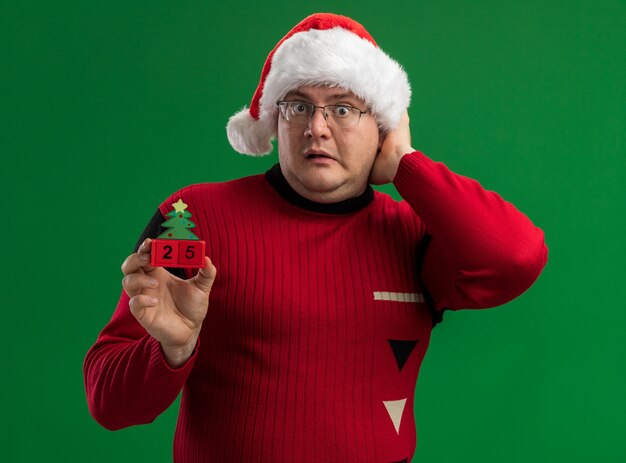 Hombre adulto preocupado con gafas y gorro de Papá Noel sosteniendo el juguete del árbol de navidad con fecha mirando a la cámara manteniendo la mano en la cabeza aislada sobre fondo verde