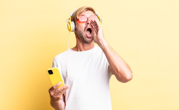 Hombre adulto rubio que se siente feliz, dando un gran grito con las manos al lado de la boca con auriculares