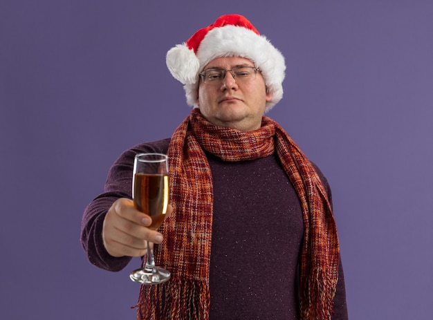 Hombre adulto seguro con gafas y gorro de Papá Noel con bufanda alrededor del cuello extendiendo una copa de champán hacia la cámara mirando a cámara aislada sobre fondo púrpura