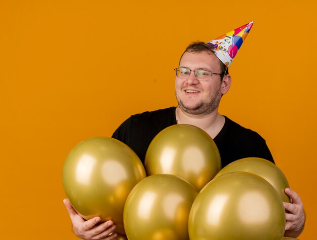 Hombre adulto sonriente con gafas ópticas con gorro de cumpleaños se encuentra con globos de helio aislados en la pared naranja