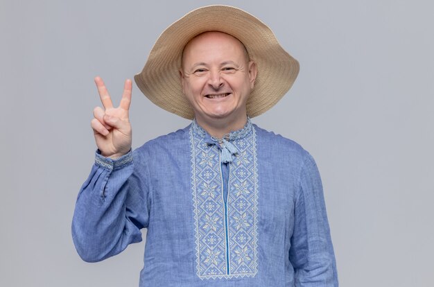 Hombre adulto sonriente con sombrero de paja y camisa azul gesticulando el signo de la victoria