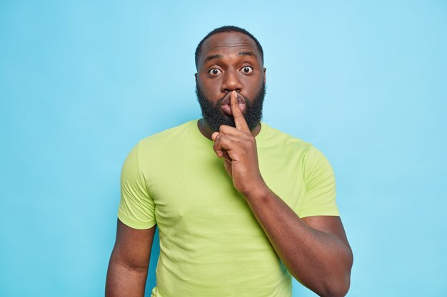 El hombre adulto sorprendido dice que shh pide estar en silencio y se sorprende al escuchar los rumores presiona el dedo en los labios usa una camiseta verde aislada sobre una pared azul