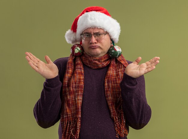 Hombre adulto tonto con gafas y gorro de Papá Noel con bufanda alrededor del cuello mostrando las manos vacías con adornos navideños colgando de sus orejas aisladas en la pared verde oliva