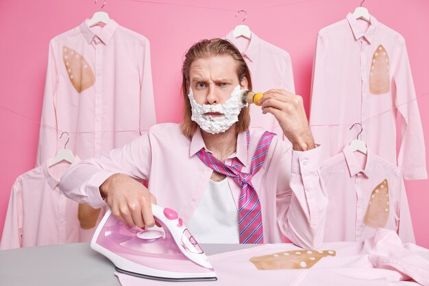 el hombre se afeita la barba se ve con expresión de disgusto usa camisa y corbata alrededor del cuello usa plancha eléctrica de vapor para acariciar puestos de ropa cerca de la tabla de planchar