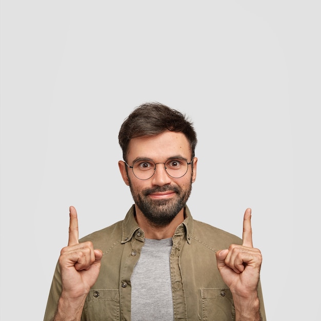 Foto gratuita hombre sin afeitar complacido de aspecto agradable nota algo asombroso hacia arriba, puntos por encima de la cabeza