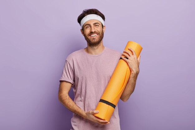 Hombre sin afeitar complacido sostiene una estera de fitness enrollada, satisfecho después de la clase de yoga