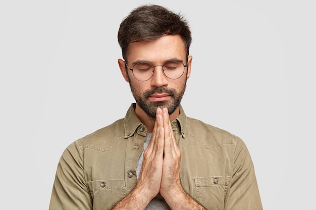 Foto gratuita el hombre sin afeitar concentrado se para en gesto de oración, mantiene las palmas juntas