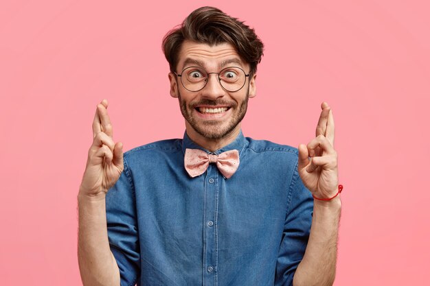 Hombre sin afeitar positivo con expresión feliz cruza los dedos, espera buena suerte, vestido con una camisa de mezclilla de moda con pajarita rosa, tiene un aspecto positivo, aislado sobre la pared