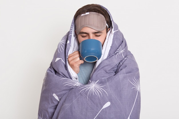 Foto gratuita hombre sin afeitar que pasa tiempo en casa por la mañana, el hombre envuelto en una manta que sostiene la taza azul con bebida caliente, disfrutando de tomar café