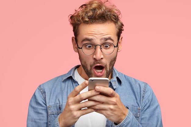 Hombre sin afeitar sorprendido mira sorprendentemente y con expresión aterrorizada en la pantalla del teléfono inteligente, usa wifi gratuito para enviar mensajes en las redes sociales, lee noticias horribles a través de Internet. Concepto de Omg
