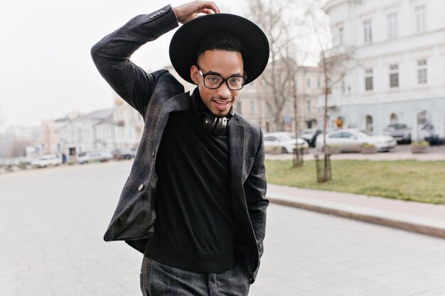 Hombre africano alegre en ropa casual caminando en el parque con auriculares. Interesado chico negro con sombrero pasar la mañana al aire libre y disfrutar de la vida.