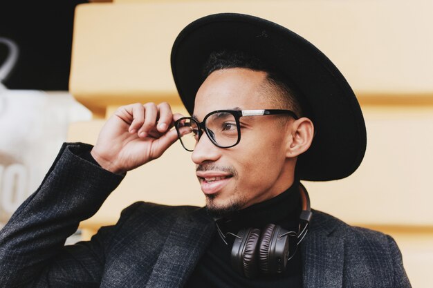 Hombre africano barbudo con corte de pelo corto tocando sus gafas. Foto al aire libre de primer plano de niño negro con sombrero y auriculares de música.