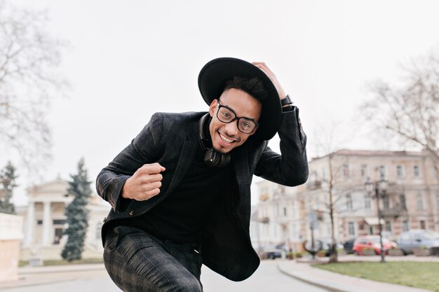 Hombre africano bien vestido bailando en la plaza de la ciudad con una sonrisa. Riendo chico negro tocando su sombrero mientras posa al aire libre bajo un cielo gris.