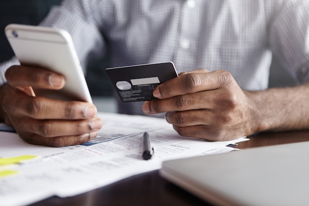 Hombre africano en camisa pagando bienes en internet con tarjeta de crédito y teléfono celular