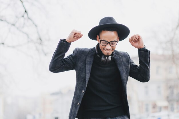 Hombre africano complacido con una sonrisa suave posando con las manos en alto. Retrato al aire libre del modelo masculino negro feliz viste traje de moda.