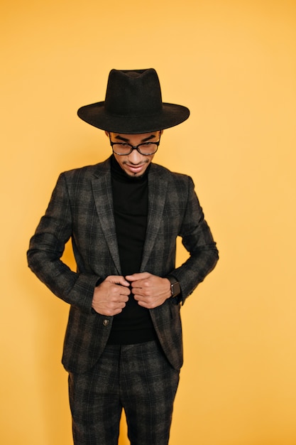 Foto gratuita hombre africano delgado con sombrero negro grande posando en traje elegante. foto interior de alegre mulato en traje gris aislado en la pared amarilla.