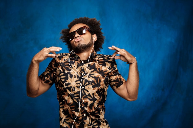 Hombre africano escuchando música en auriculares, bailando sobre la pared azul.