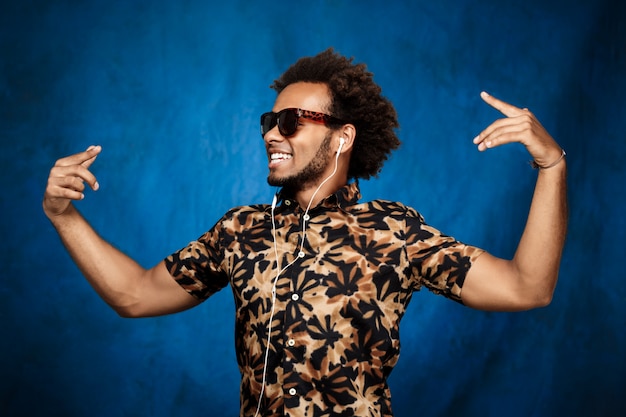 Hombre africano escuchando música en auriculares, bailando sobre la pared azul.